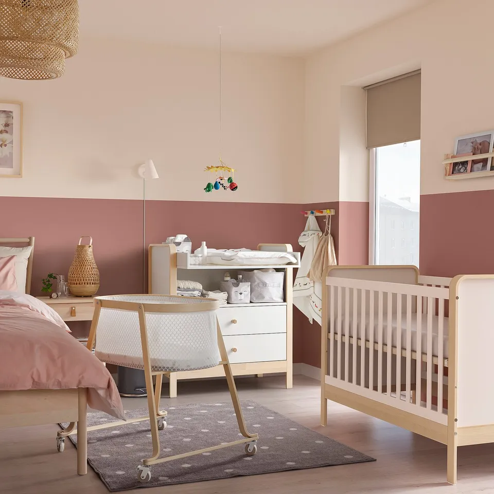 Babys room with white ÄLSKVÄRD bassinet, changing table, light pink walls, wooden floors, and polka dot rug.
