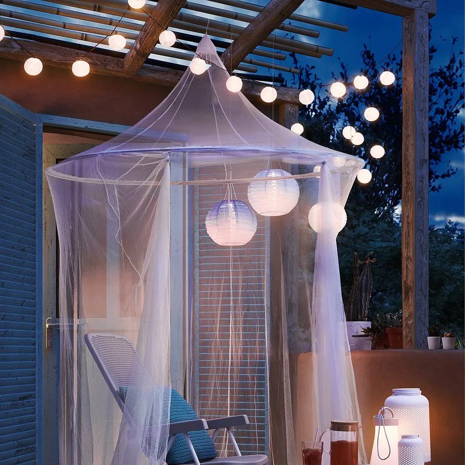 Relaxing patio scene with a white SOLIG net canopy, string lights, blue chairs, and table holding drinks and lanterns.