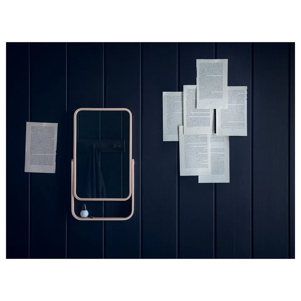 Ikorennes mirror hangs on dark wall, framed in light wood with equivalent stand, reflecting partially visible room. Papers stuck beside and around it.