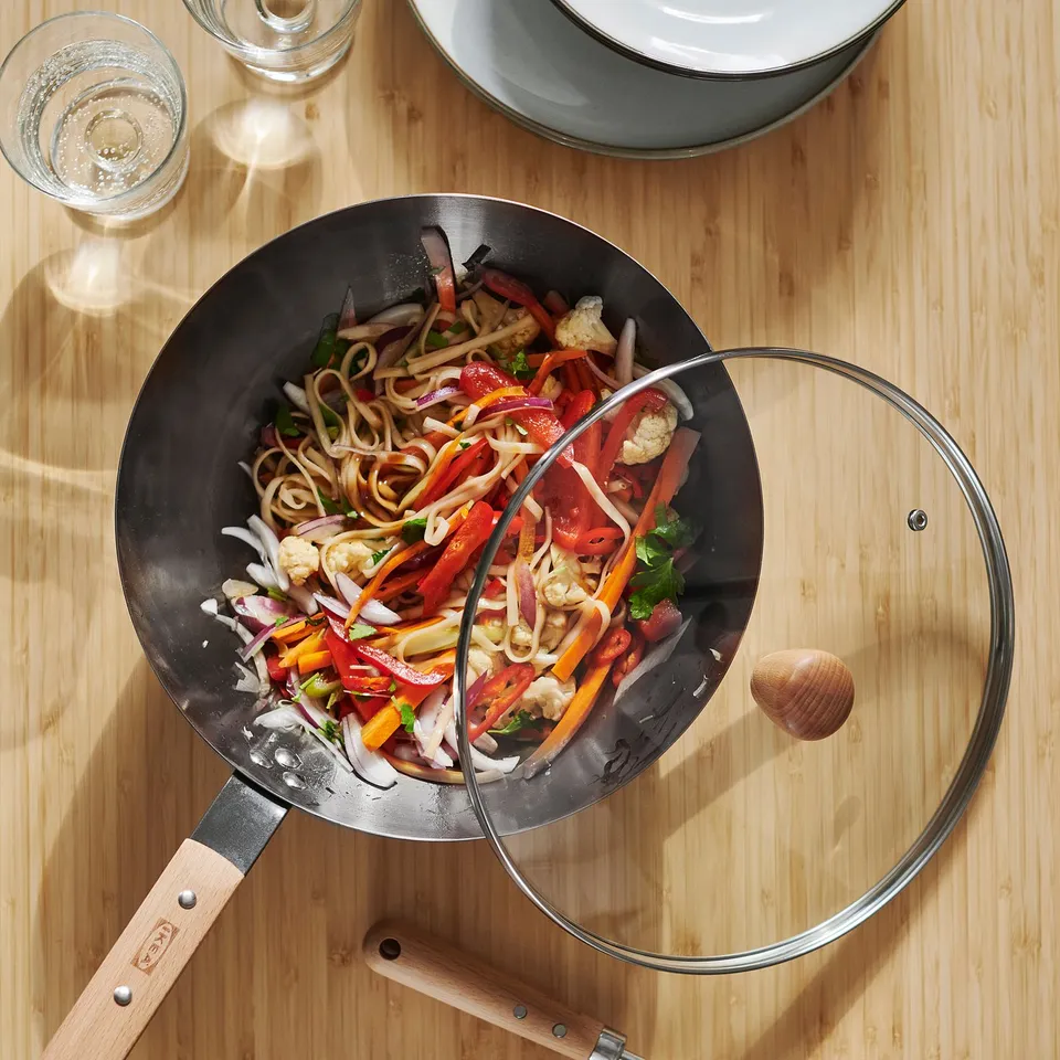 Top-down view of a wok on a wooden table, filled with stir-fried vegetables and noodles. The wok has a glass lid and wooden handles.