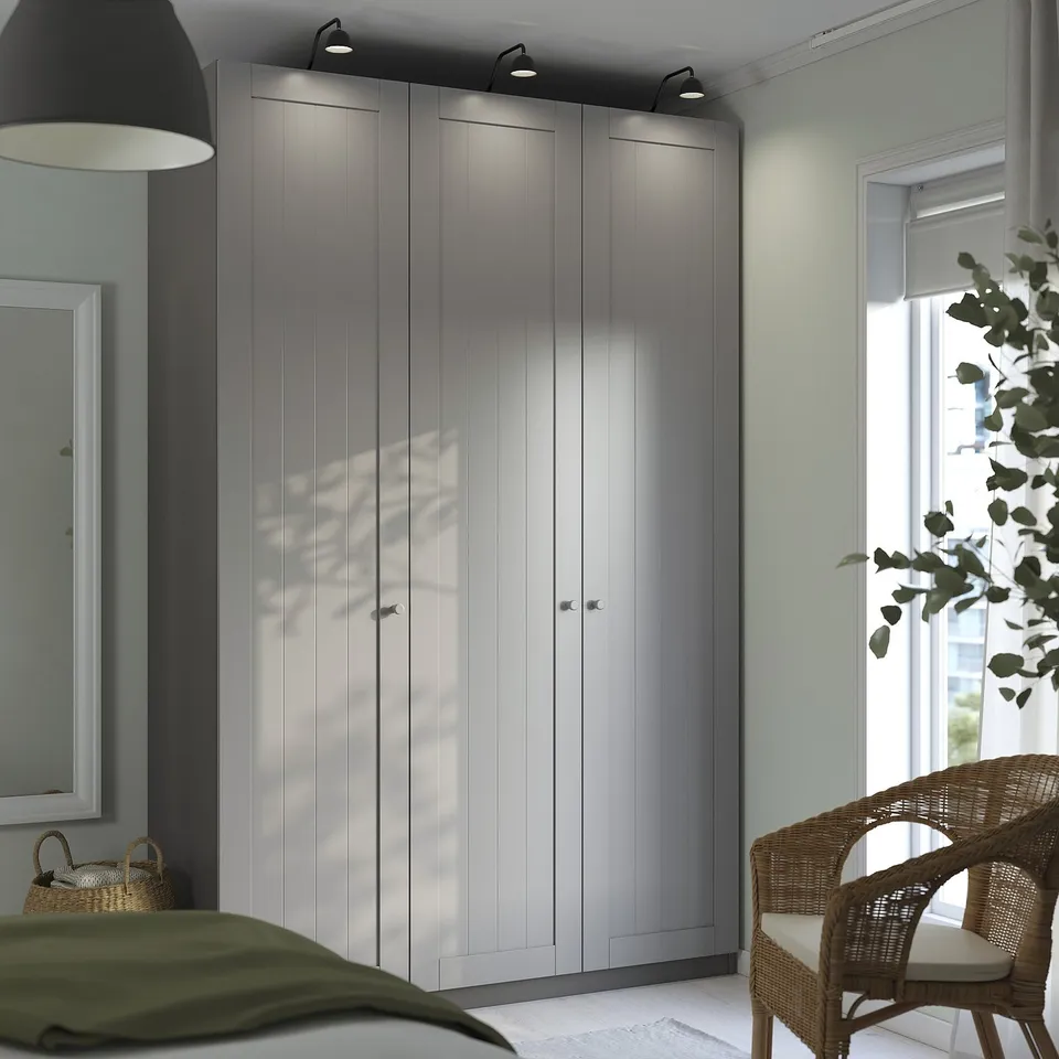 Modern bedroom with grey PAX wardrobe, wall lamp, window, wicker chair, and green bedspread.