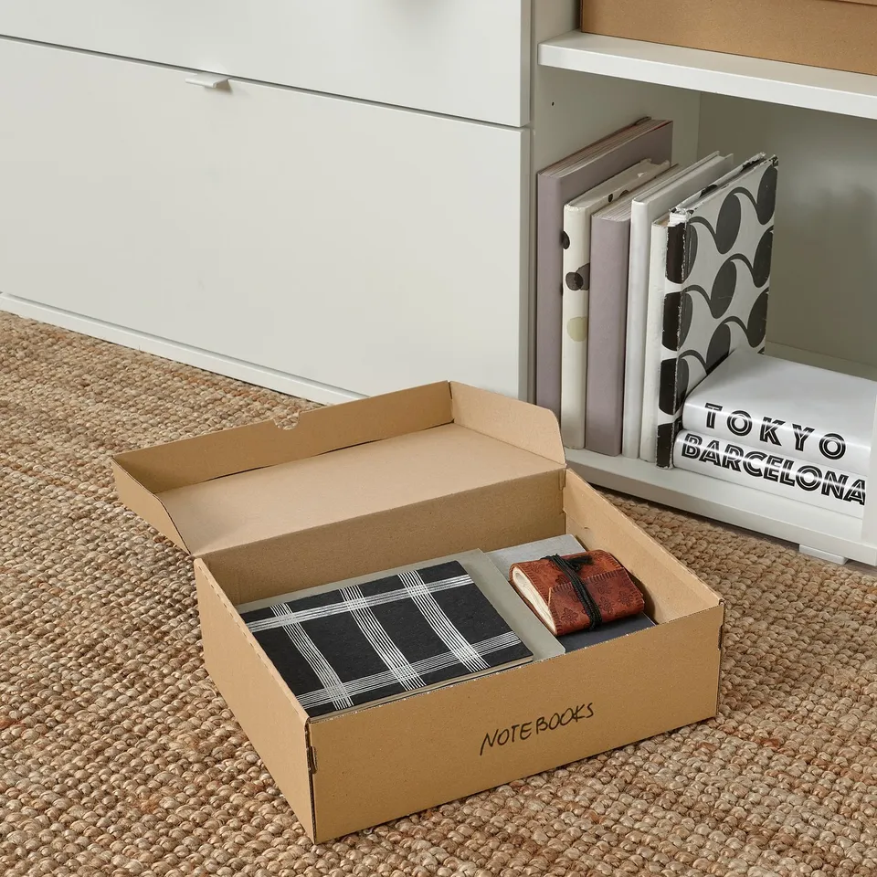 An open cardboard box with note books and a pocketbook on a jute rug, labelled note books.