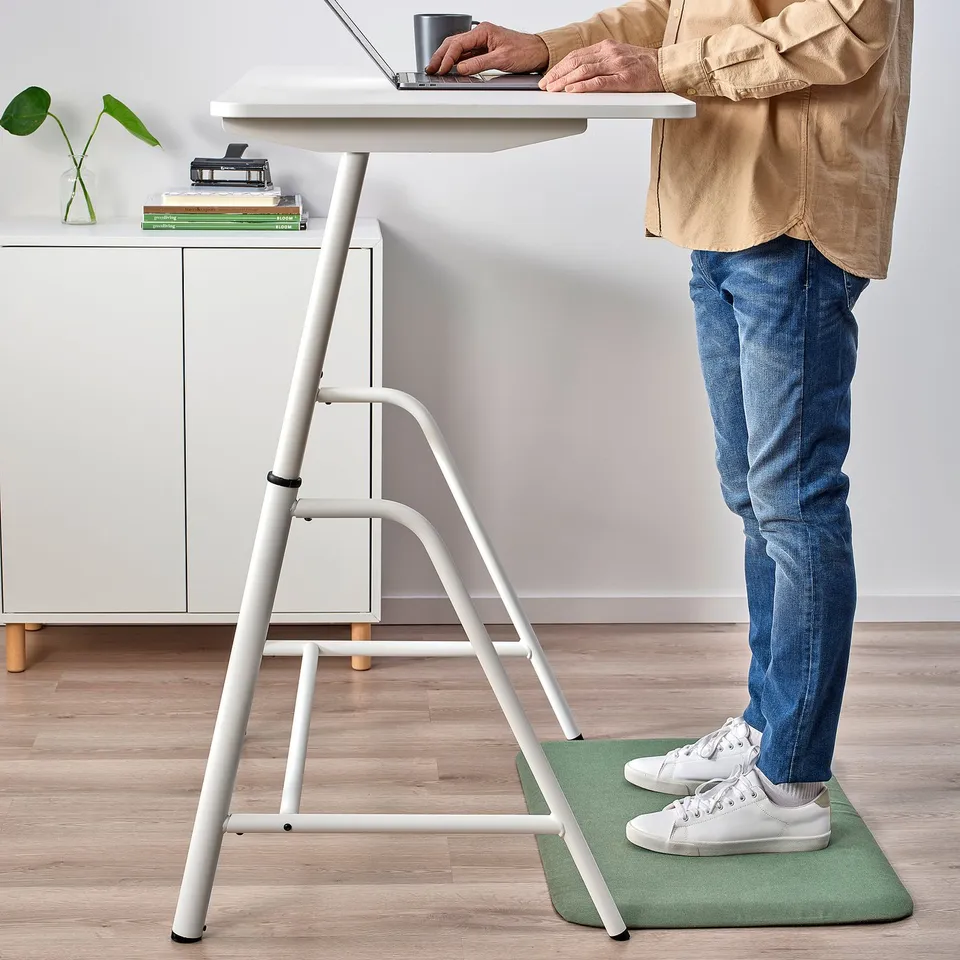 Person stands on green mat while using laptop at high desk.