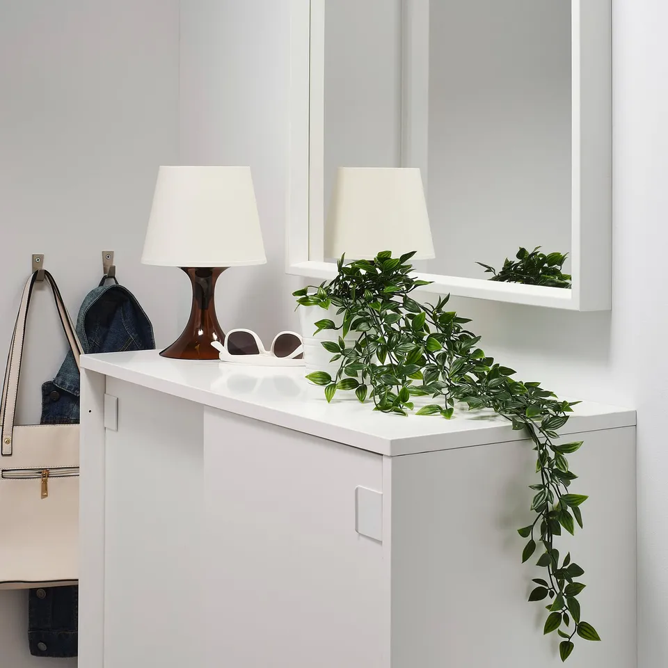 A modern interior featuring white lamps on top of a white cabinet, with FEJKA green artificial plants decorating the space. Handbags hang nearby.