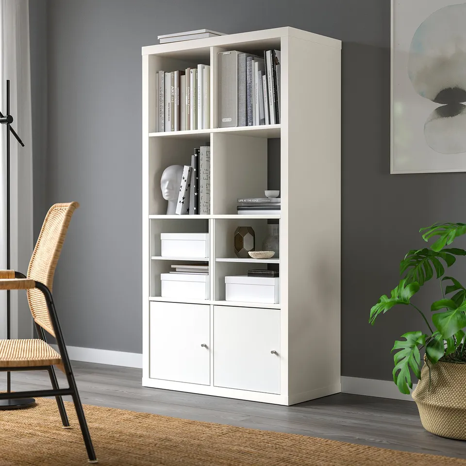 A KALLAX white bookshelf against a dark wall, filled with books and decor. It stands tall compared to a wicker chair beside it.