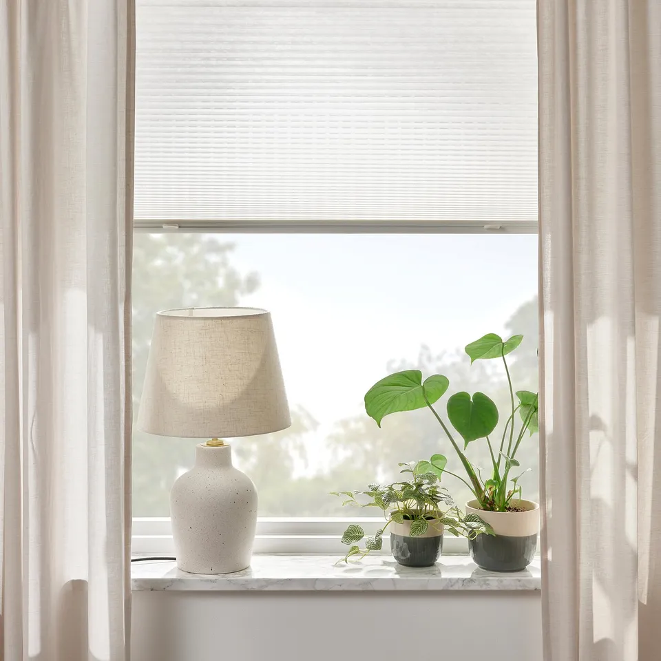A window with light-filtering pleated blinds and a marble shelf holding a lamp and plants.