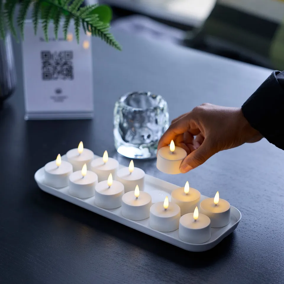 Hand picks lit tealight from tray of 12, warm ambiance. Near crystal holder, qr sign.