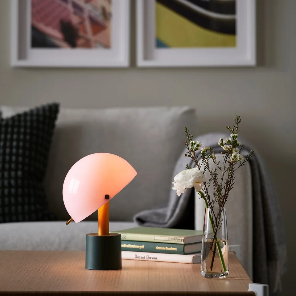 Small, modern NÖDMAST lamp in orange sits on table next to flower vase; soft light, stylish design.