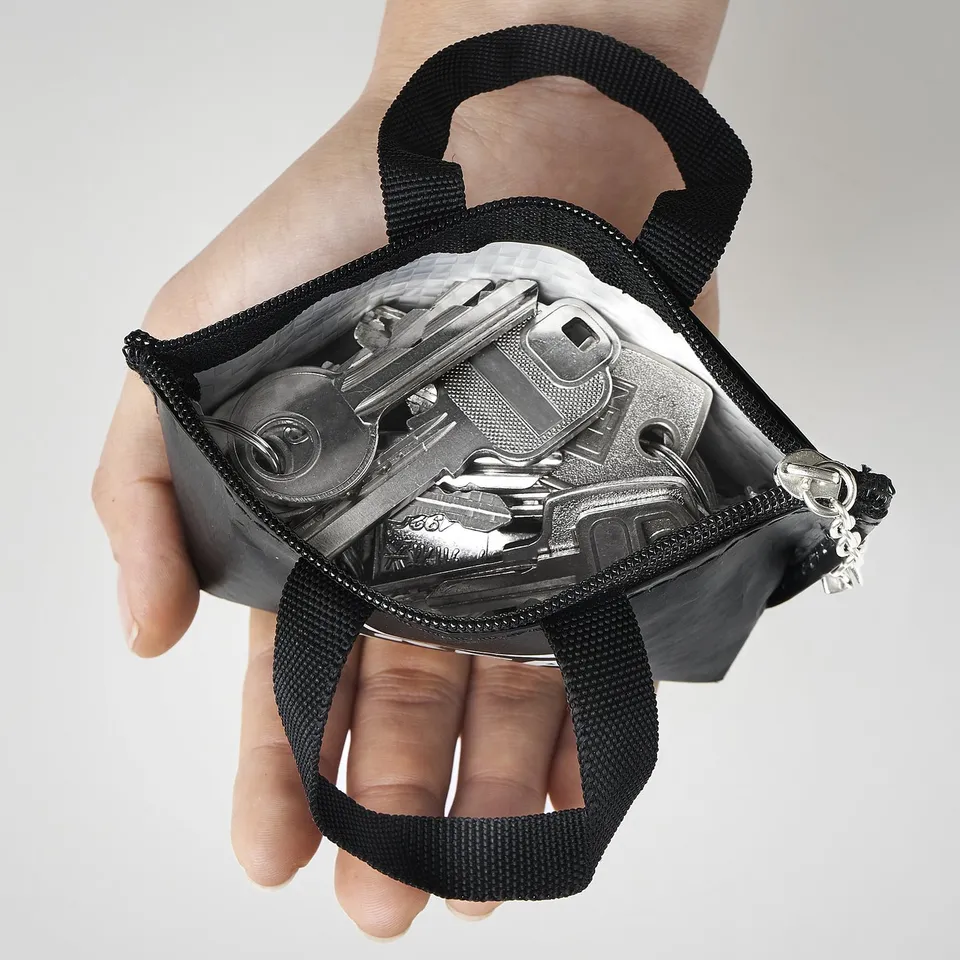 Hand holds small black KNÖLIG bag, open showing coins and keys inside, with black strap.