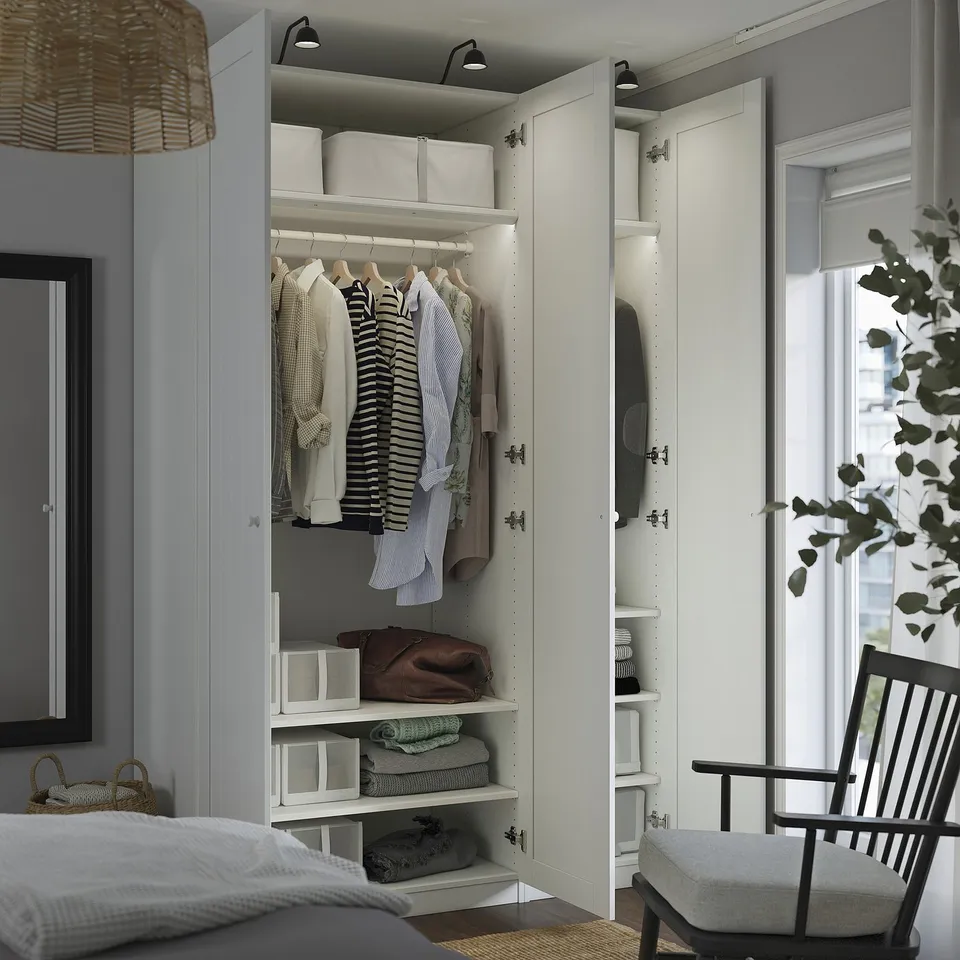 Open closet with shirts, coats, bags, and shelves in bedroom. Ceiling lights on, set beside window.