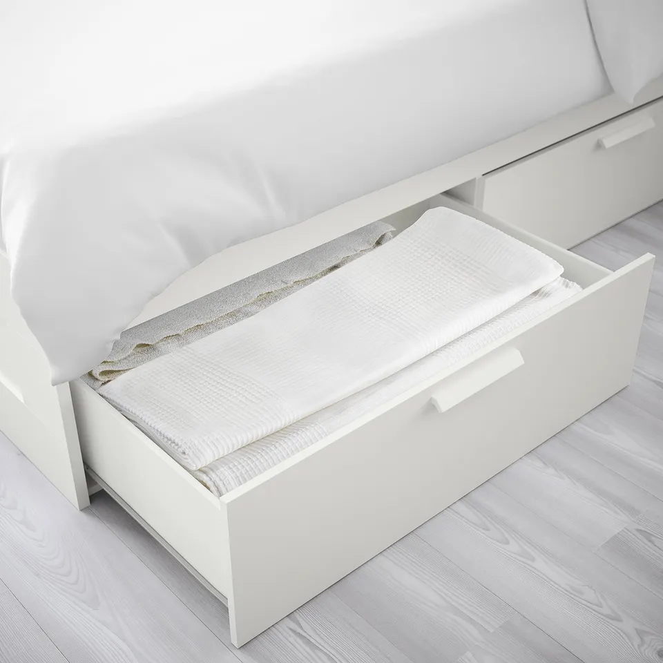 White bed frame with open drawer, showing folded bedding inside.