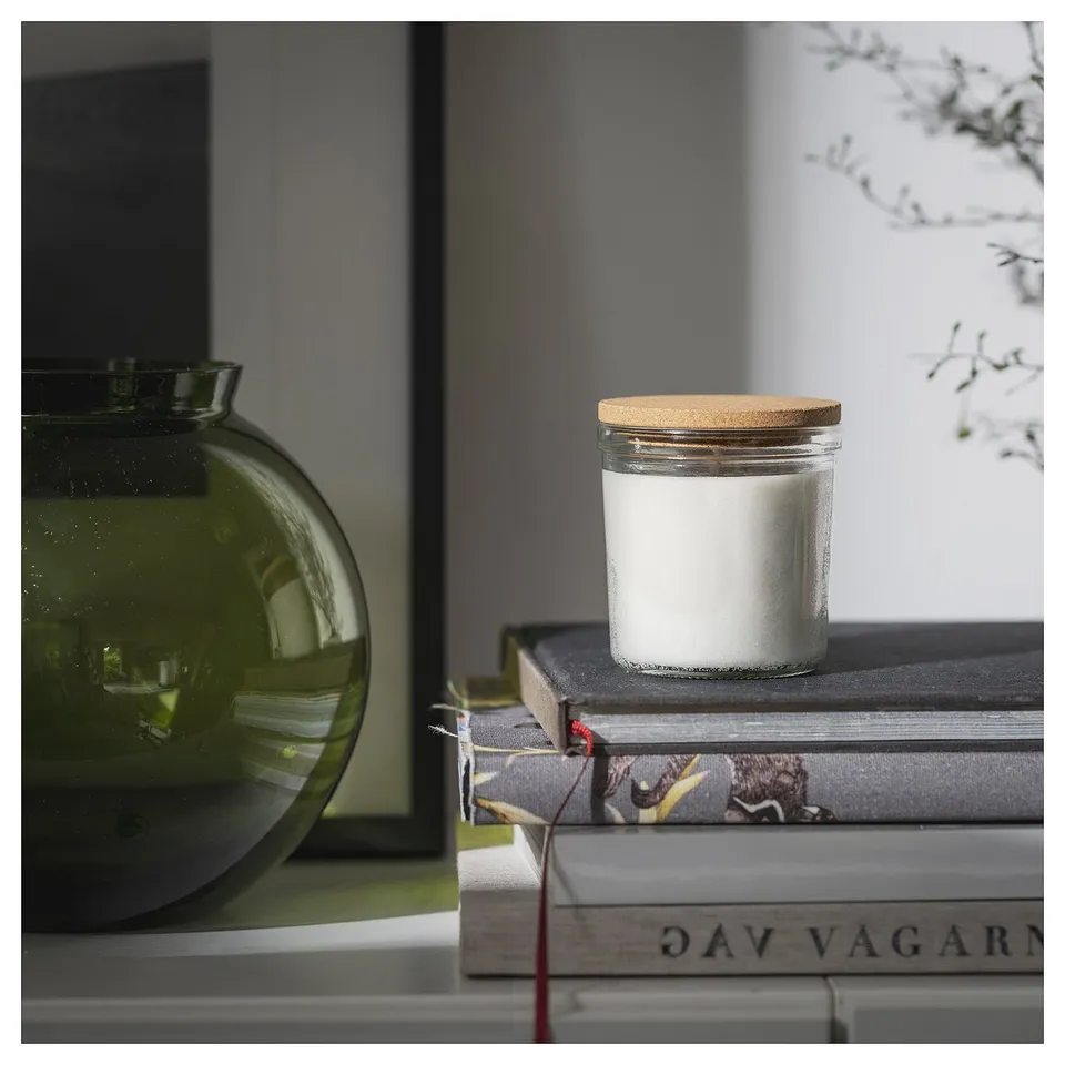 A white candle in a glass jar with a cork lid sits on stacked books next to a green vase on a shelf.