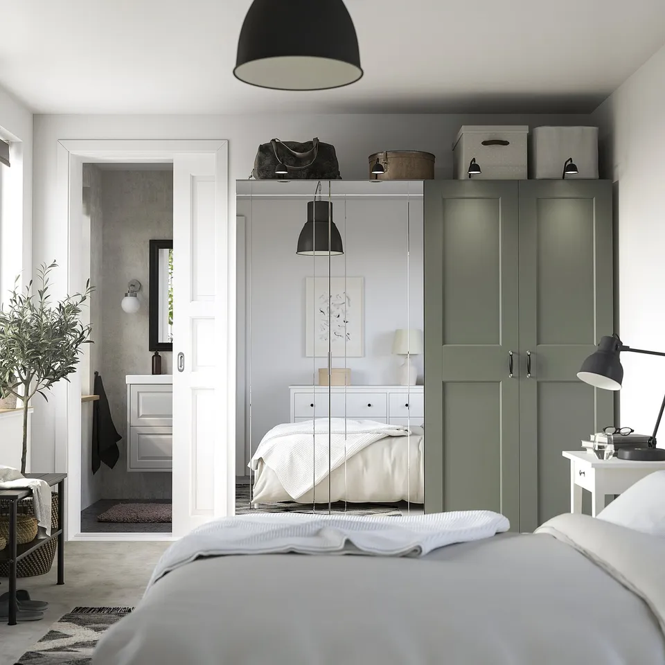 Modern living space with large bed, green PAX wardrobe, bathroom doorway, desk, lamp, and plant.