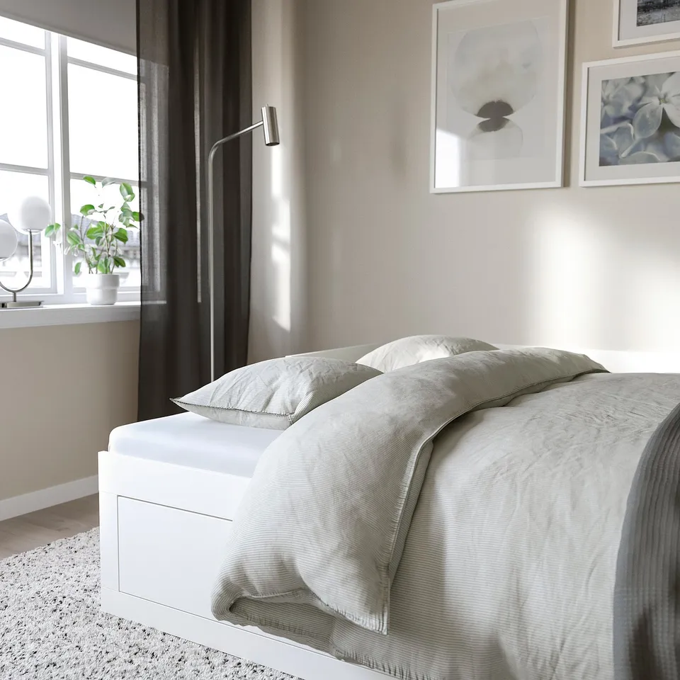 Modern bedroom with white daybed, drawers, light bedding, window with dark curtains, wall art, and plant.