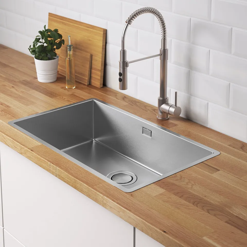 Stainless steel kitchen sink with pull-out tap, set on wooden countertop. Includes lid for sink drain and small plant.