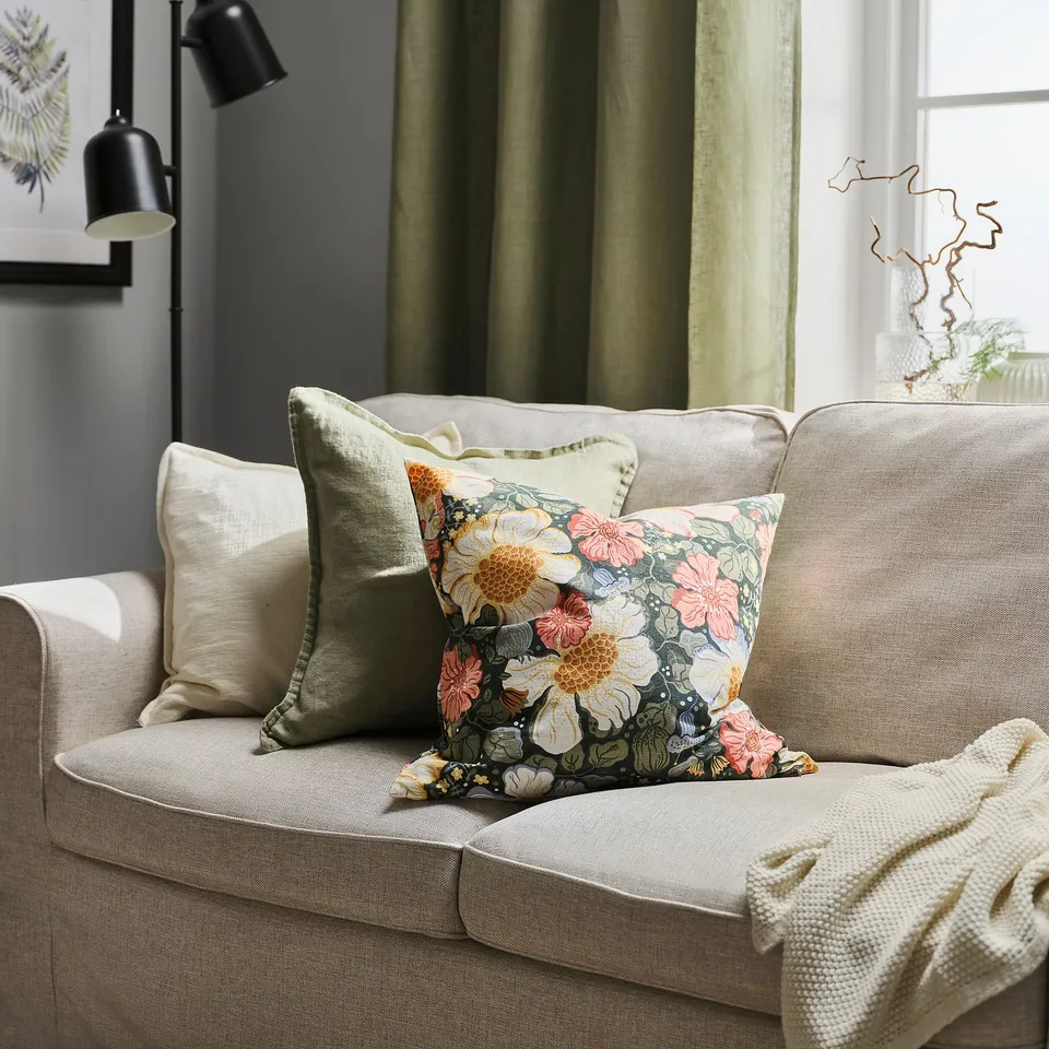 An ARENBOK cushion on a beige sofa, adorned with a vibrant floral print, surrounded by other cushions, curtains, and a lamp.