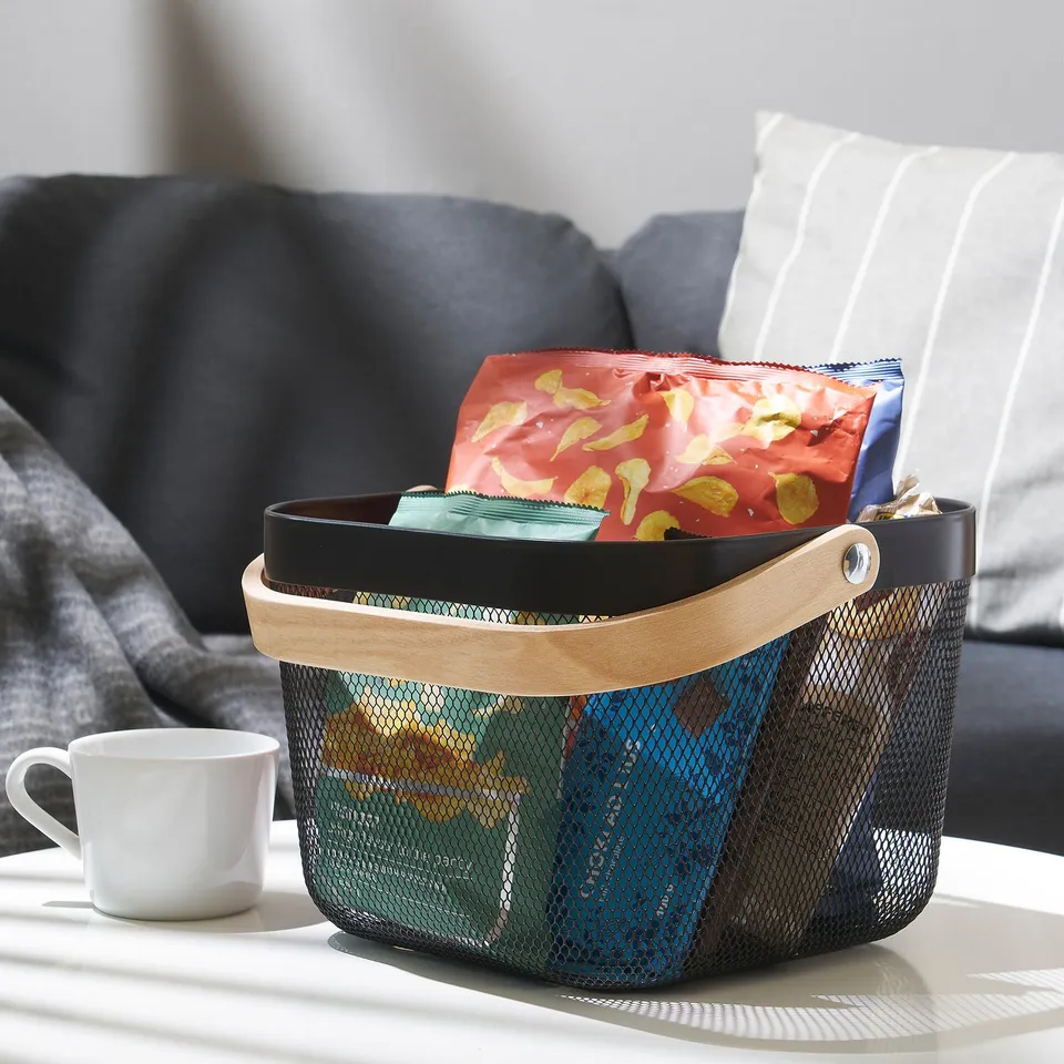 Mesh basket with wooden handle on table, holding snacks near a grey couch.