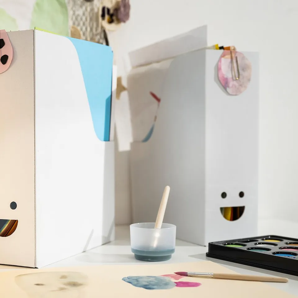 Three cheerful TJABBA magazine files with colourful smiles on a white table, holding office supplies, featuring recycled material.