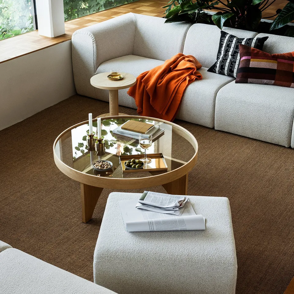 Modern living room: light grey sofa, orange throw, wooden coffee table with glass top, brown carpet, natural light, plants.