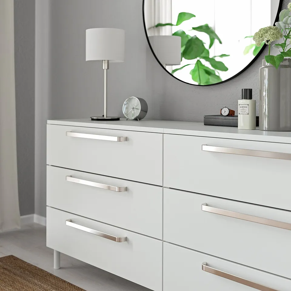 White dresser with six drawers, silver handles, round mirror, lamp, clock, and plant on top.