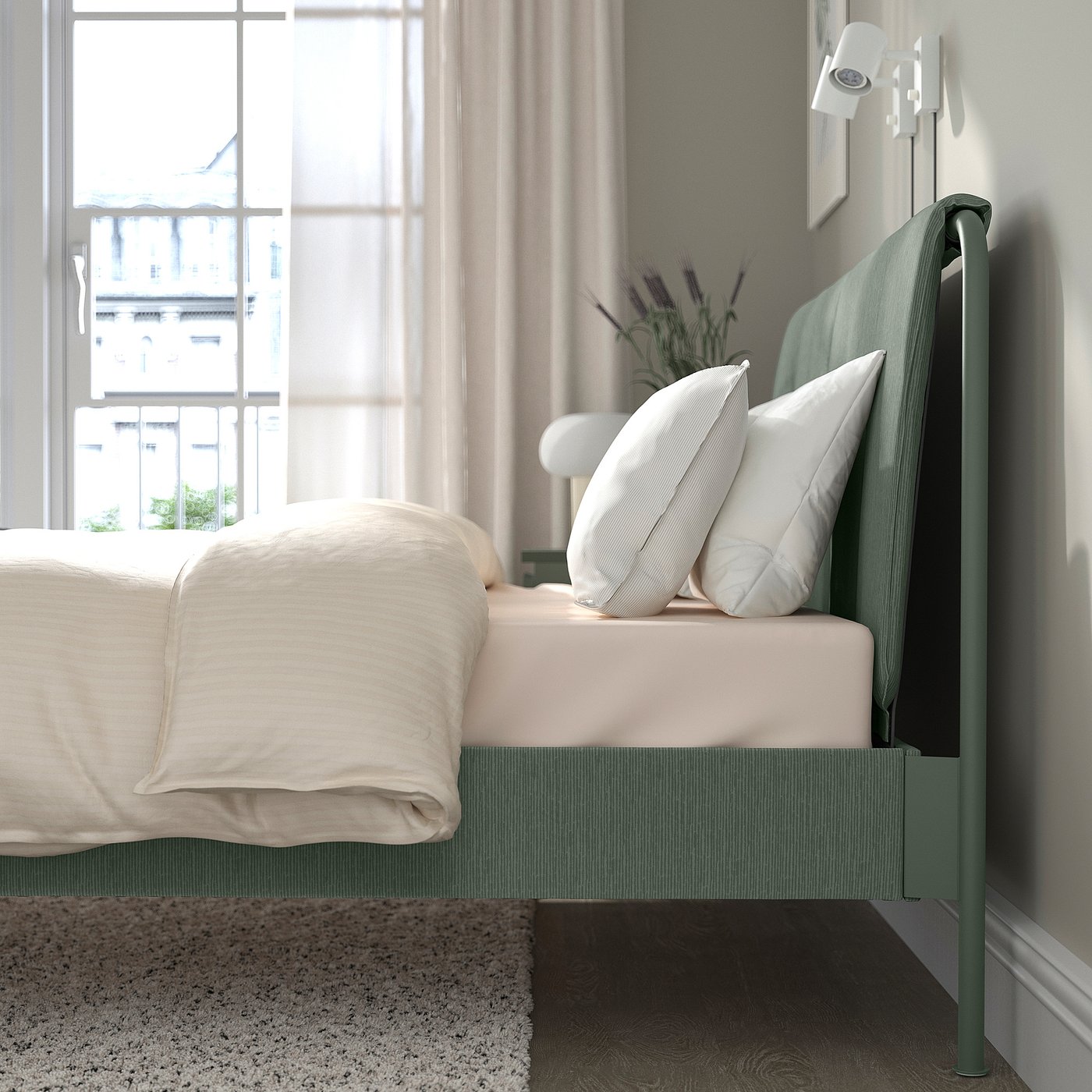 Modern green bed by window, white linens, pillows, lamp and vase with flowers on headboard.