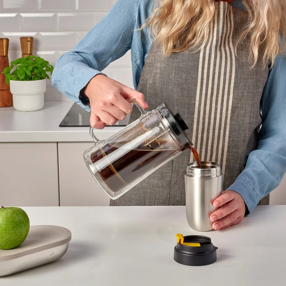 Person pouring coffee from clear pitcher into insulated, stainless steel travel mug with leak-proof lid on counter.