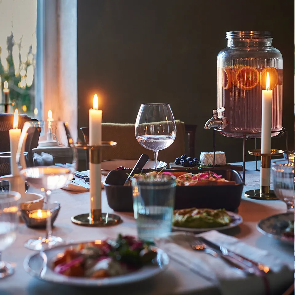 Elegant dining table set with plates, glasses, candles, and a clear jar dispenser. Soft lighting from lamps overhead creates a cosy ambiance.