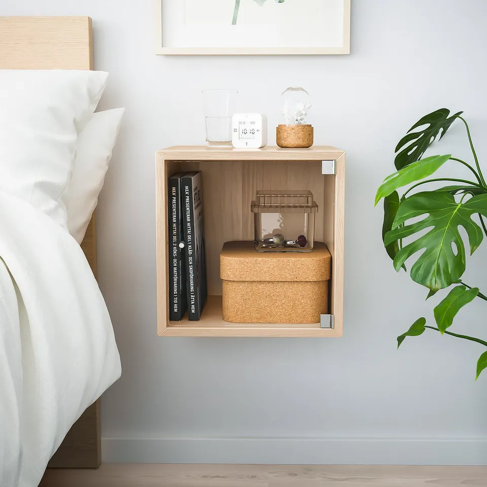 Small, wooden, wall-mounted cube shelf holds books, boxes, and decor. Next to white bedding and large green plant.