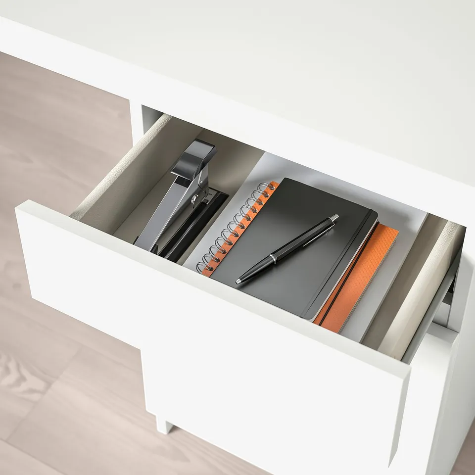 White desk drawer open, revealing a pen, notebook, and stapler, showing organised workspace storage.