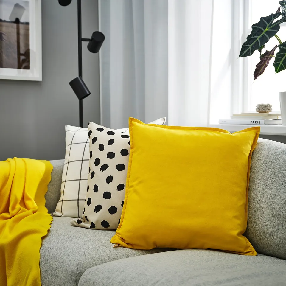 Bright yellow GURLI cushion, square with zip, on grey sofa. Beside patterned pillows, near window with plant.