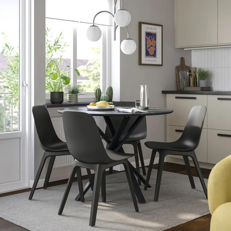 Modern dining setup with black chairs & table, sleek designs, fruit plate & vase, plants.