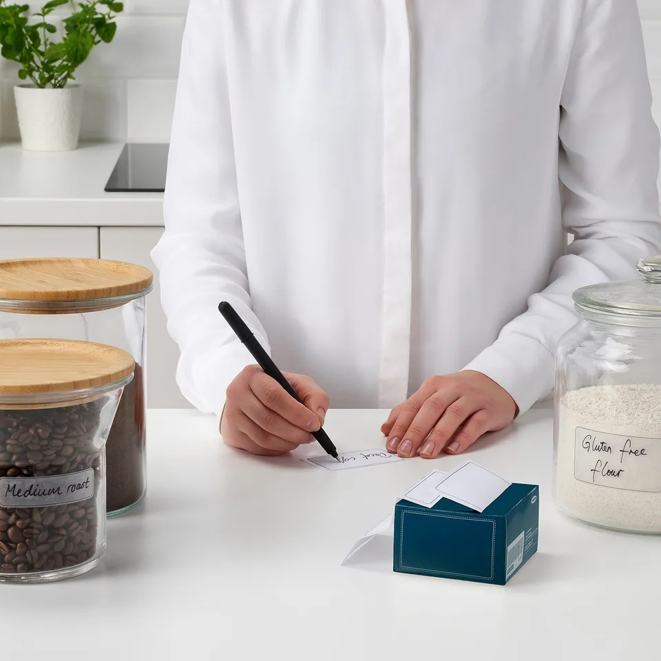 Person writes on label near food jars. Jars labelled medium roast and gluten free flour, dishwasher-safe labels.