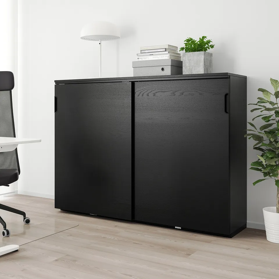 Black GALANT sliding cabinet in office, beside desk, with books and plant.