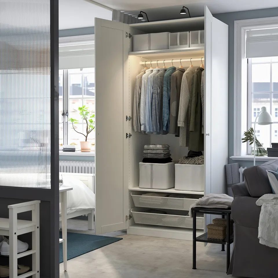 Modern bedroom with open PAX wardrobe displaying clothes, storage bins, and integrated lighting.