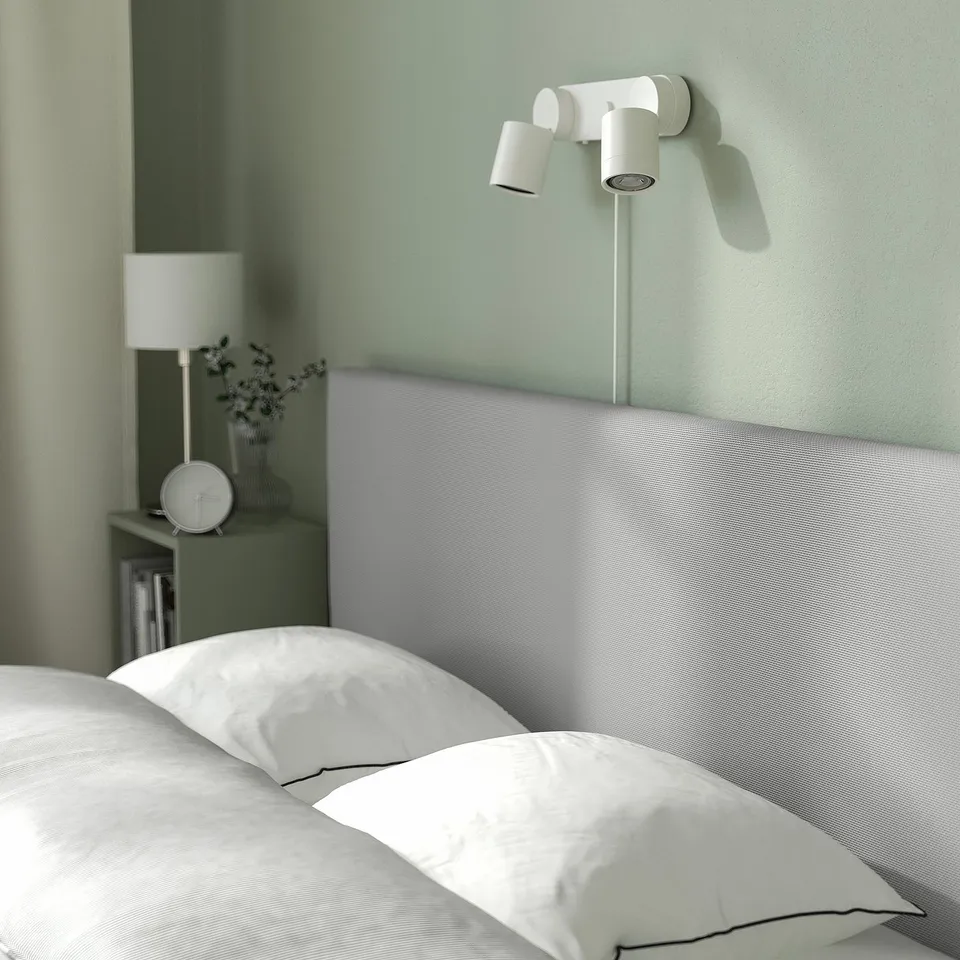 Modern bedroom with GLADSTAD bed showing wall lamps, neatly made white bedding, lamp, clock, and small plant on bedside table.