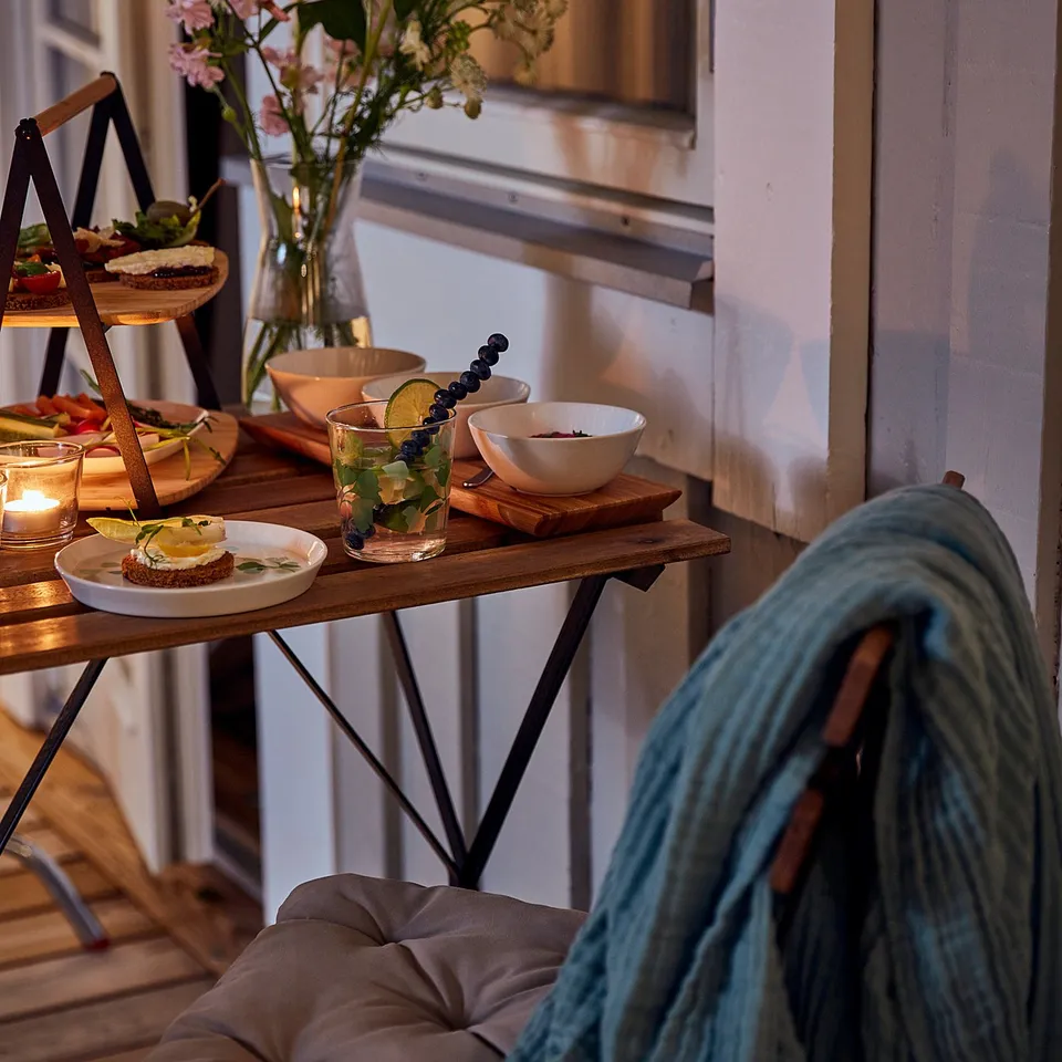 Cosy balcony scene with TÄRNÖ brown foldable furniture set. Beverages, snacks, and flowers on the table. Person in blue blanket seated nearby.