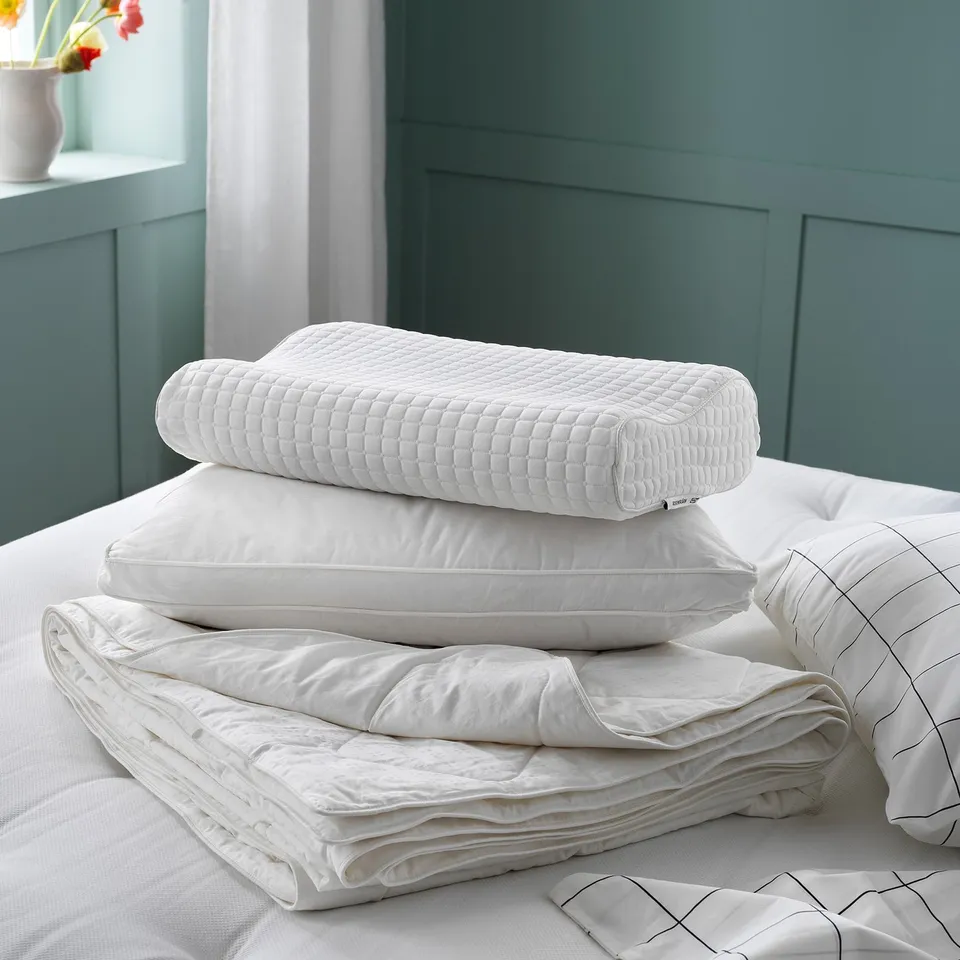 White bedding set with green walls; includes square-patterned pillow, flat pillow, folded white blanket, and striped decorative pillow.