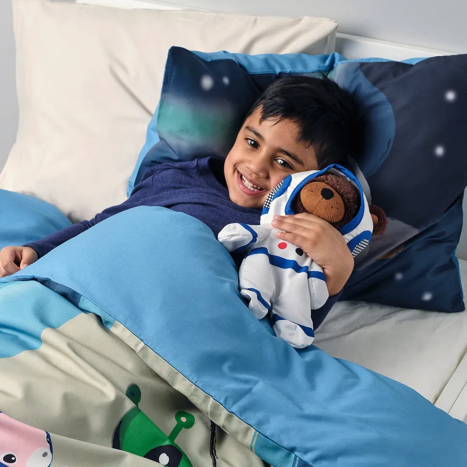 In a kid smiles while holding an astronaut teddy bear, lying comfortably on a space-themed bed with pillows and blankets.