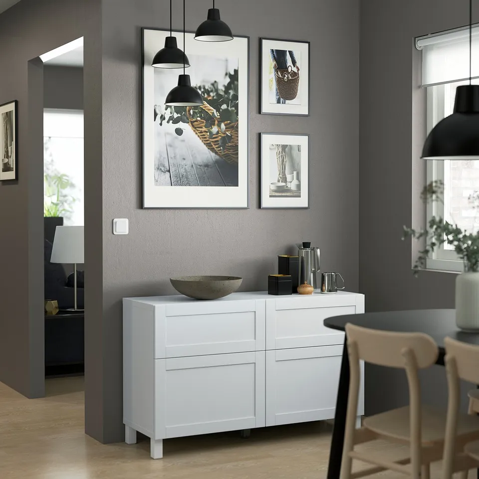 A BESTÅ storage unit with drawers and shelves against a grey wall, featuring art, lamps, and a dining setup.