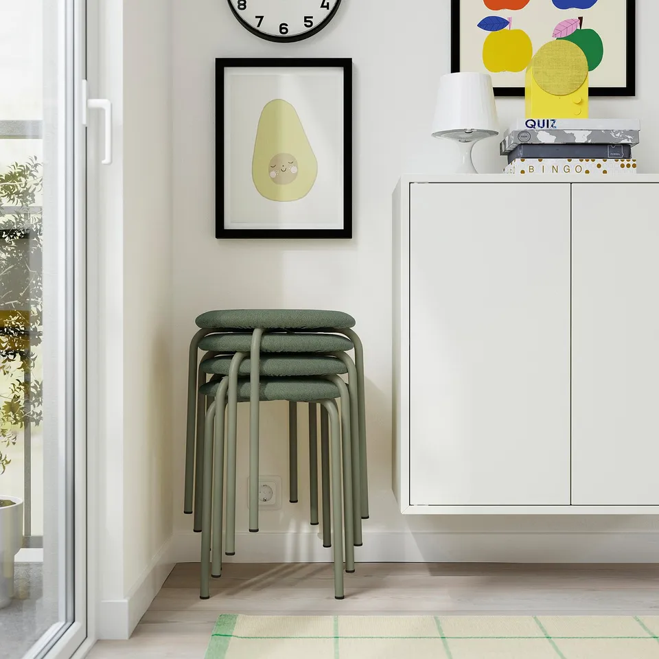 A modern, minimalist room with a stack of green stools, a clock, a plant, colourful art, and a white cabinet with games on top.