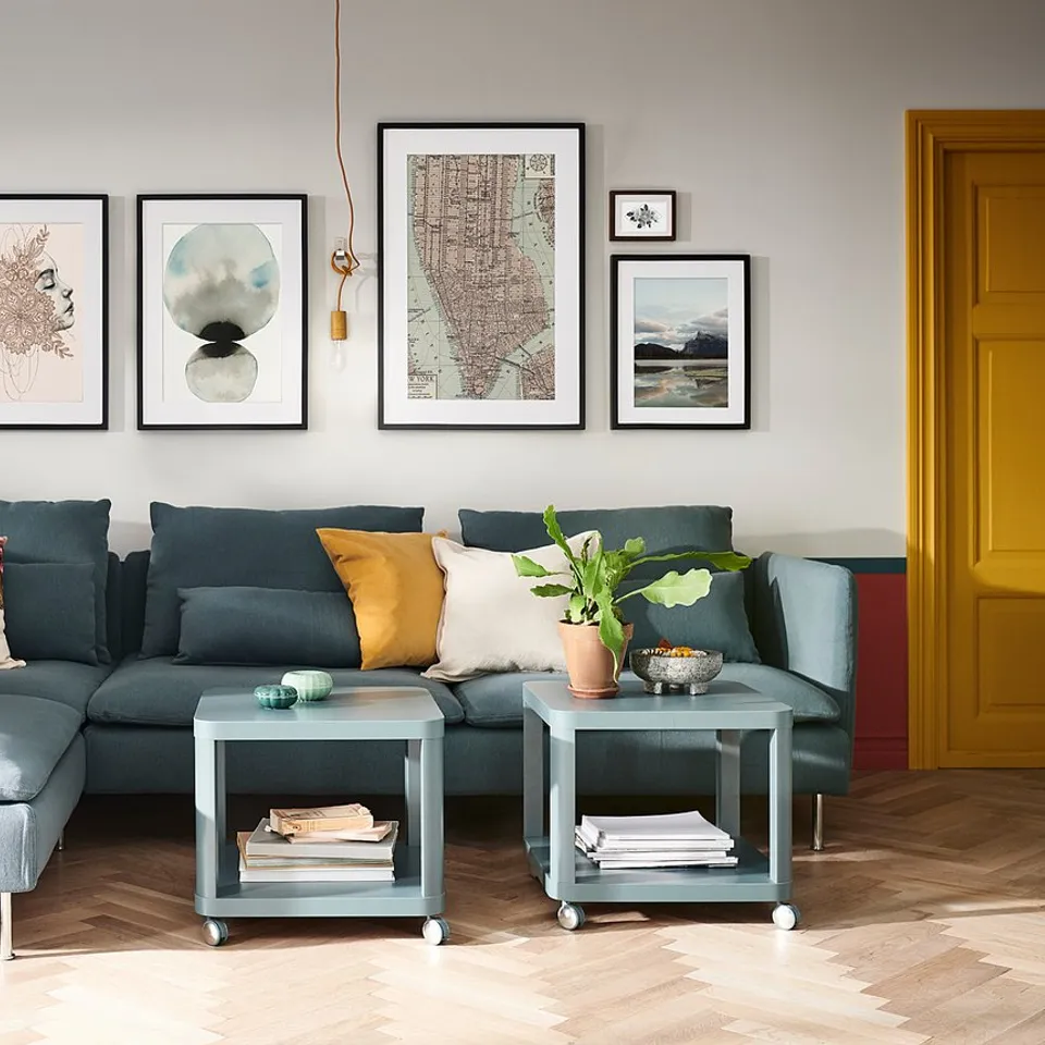 Modern living room with teal sofa, colourful pillows, art wall, grey side tables with plants, and a large green plant.