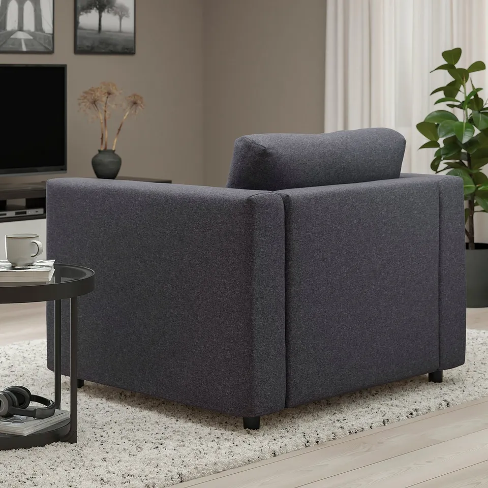 A dark grey upholstered chair in a living room, positioned near a table with a cup and remote, under a picture frame and beside a potted plant.