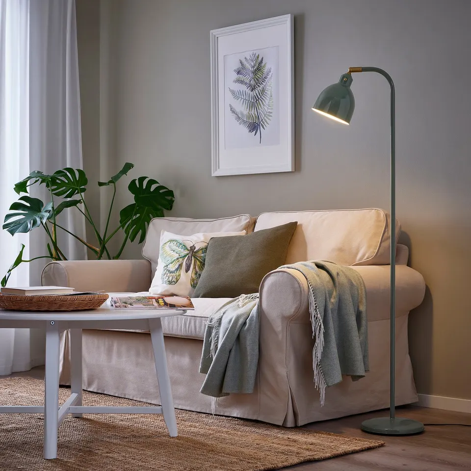 Cosy living room with beige lamp, green shade, light sofa, grey blanket, coffee table plant, and grey wall with botanical print.
