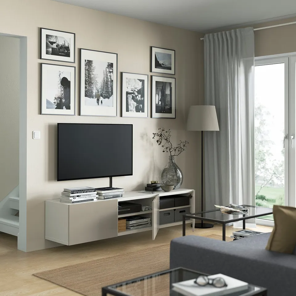 Modern living room with BESTÅ tv bench, flat-screen tv, grey sofa, glass coffee table, tall lamp, and large window with light grey curtains.