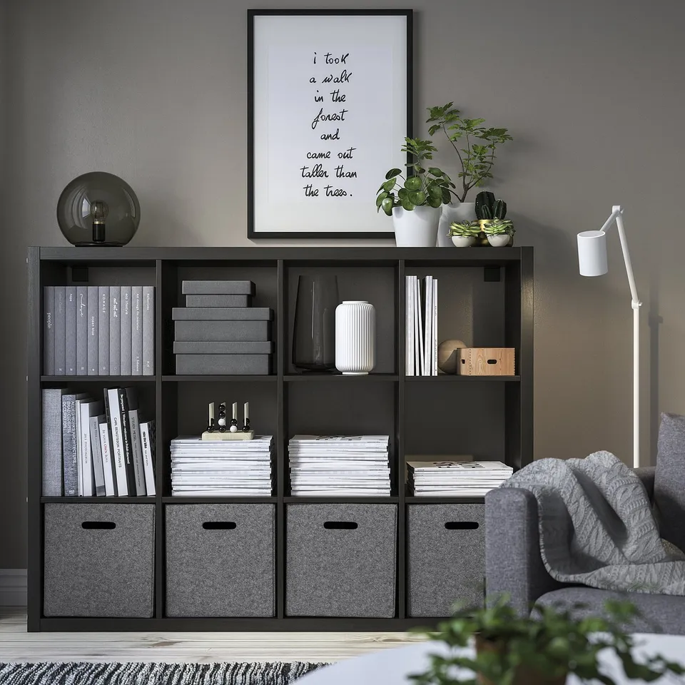 Modern room with BLÄDDRA storage boxes in kallax unit, holding books, decor, and plants. Framed quote and lamp on top shelf, with grey chair and rug.