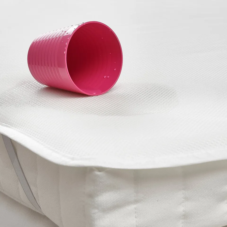 A bright pink cup rests on a white quilted mattress protector, designed to guard against spills and keep mattresses clean and dry.