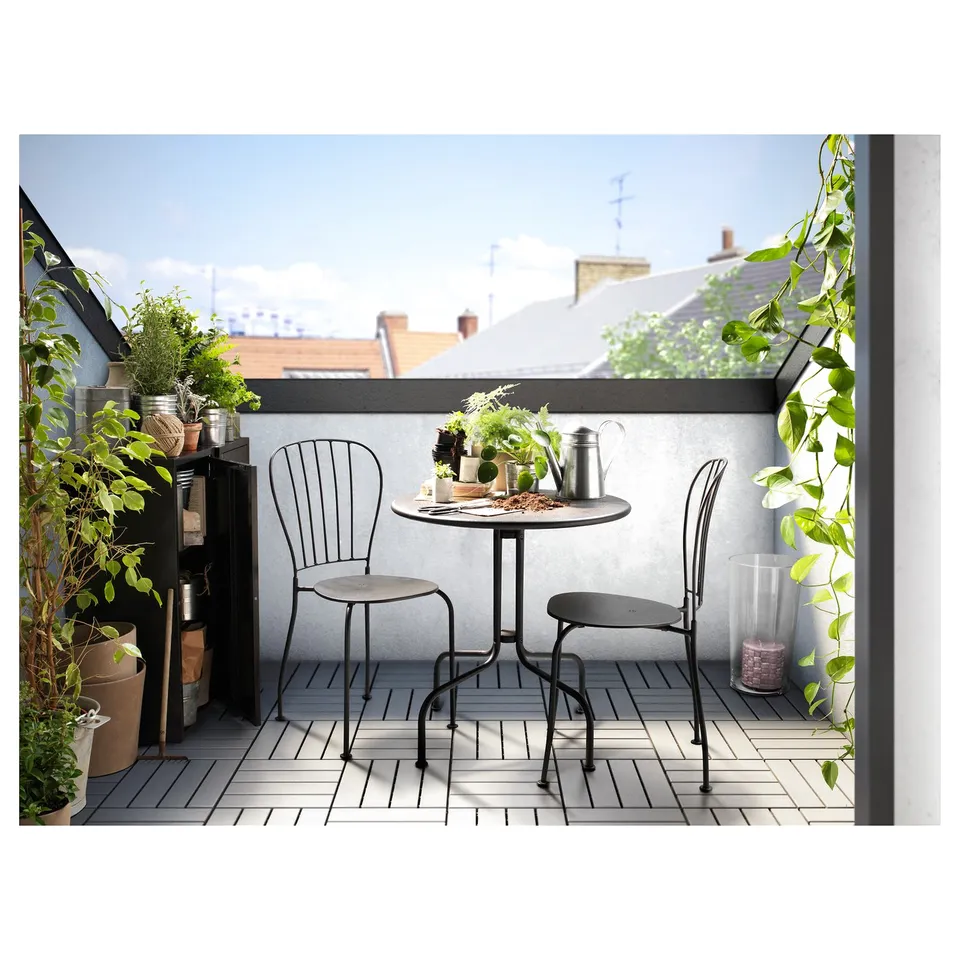 Small balcony with black metal table and two chairs, grey cushions, and potted plants.