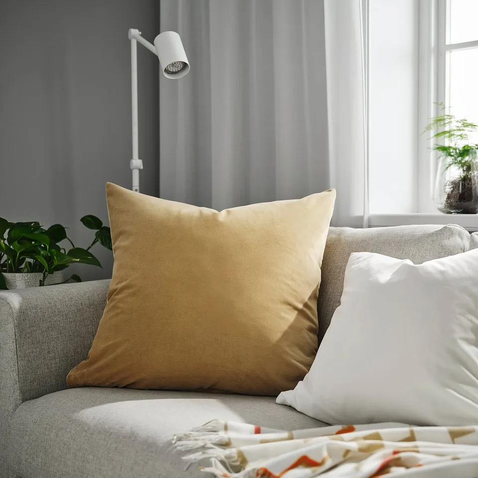 Large gold SANELA cushion on beige sofa next to white pillow, modern lamp, green plant, and bed with colourful blanket.