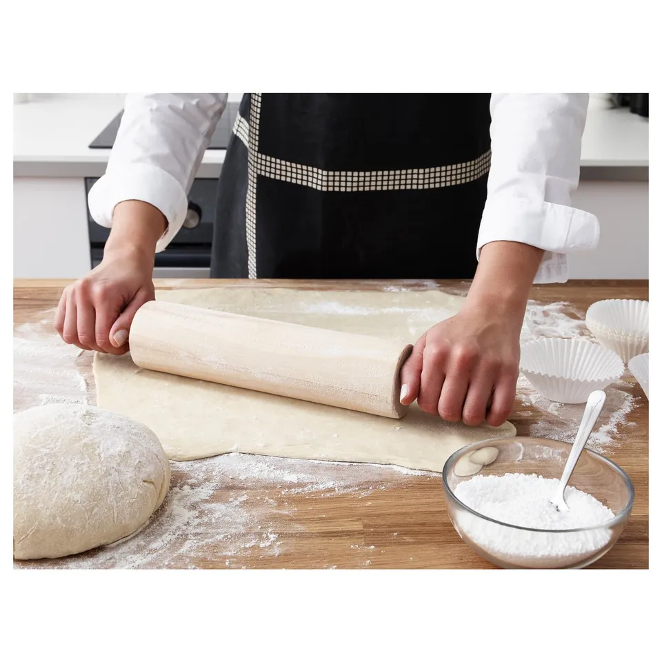 Person rolling dough with rolling pin on floured wooden table, bowl of flour nearby.