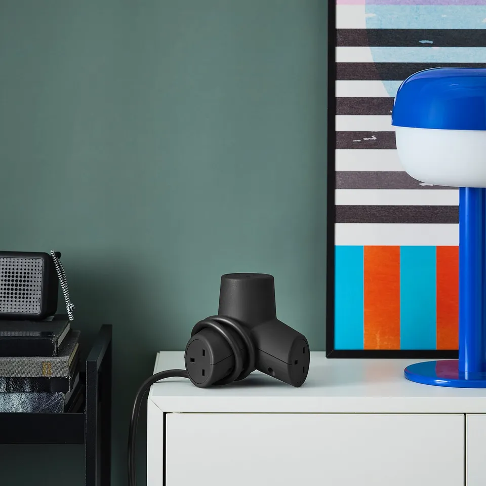 Black HORNFELS 4-way extension sits on white desk, small, near blue lamp & retro radio.